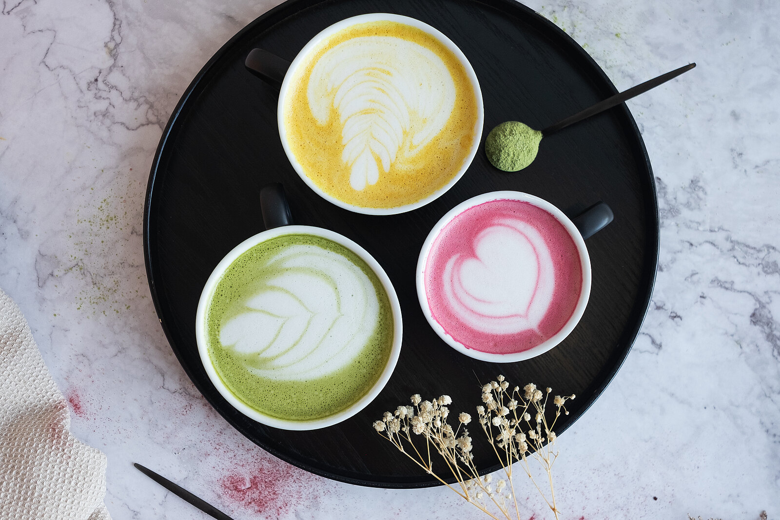 Ein Cappuccino Trio: Ein Matcha Cappuccino, ein normaler und einer mit Himbeerextrakt. Dadurch ist der Milchschaum eingefärbt in grün, gelb/braun und rot.