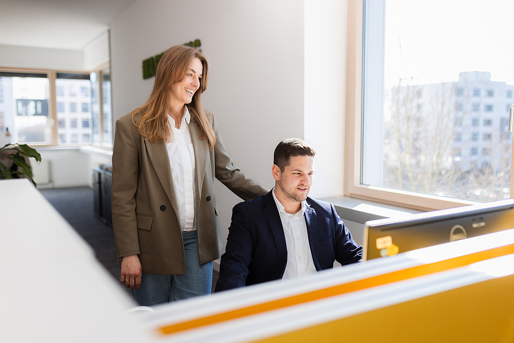Eine junge Dame, die einem jungen Herren im Büro über die Schulter blickt. Der Herr sitzt und schaut in seinen Laptop, die Dame steht neben ihm und tut das ebenso.
