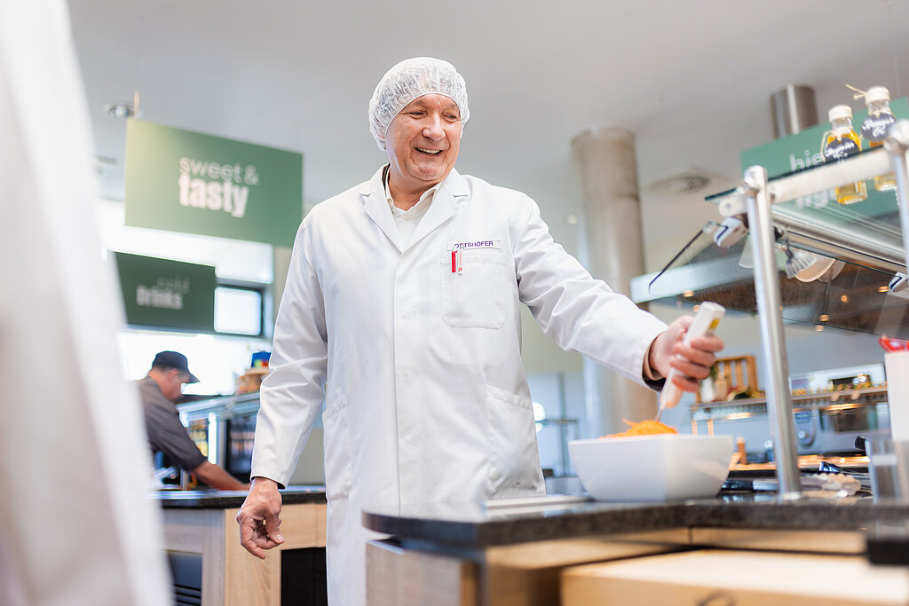 Ein Mann mit weißem Kittel und Haarnetz - er ist von der Lebensmittelkontrolle - und kontrolliert ein Salatgericht.