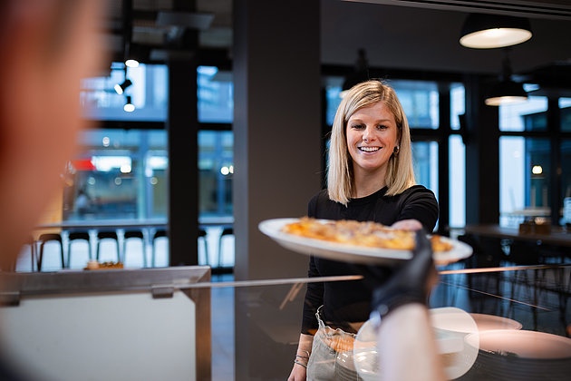 Eine junge blonde Dame, die ein Gericht entgegennimmt. Es ist eine Pizza.