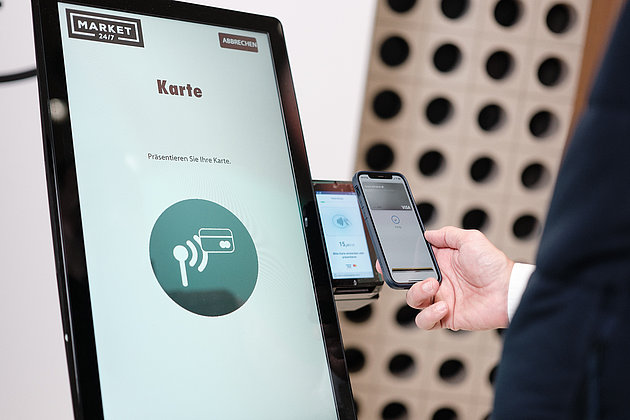 Ein Monitor, daneben eine Zahlfunktion und eine Hand die mit dem Handy bezahlt.