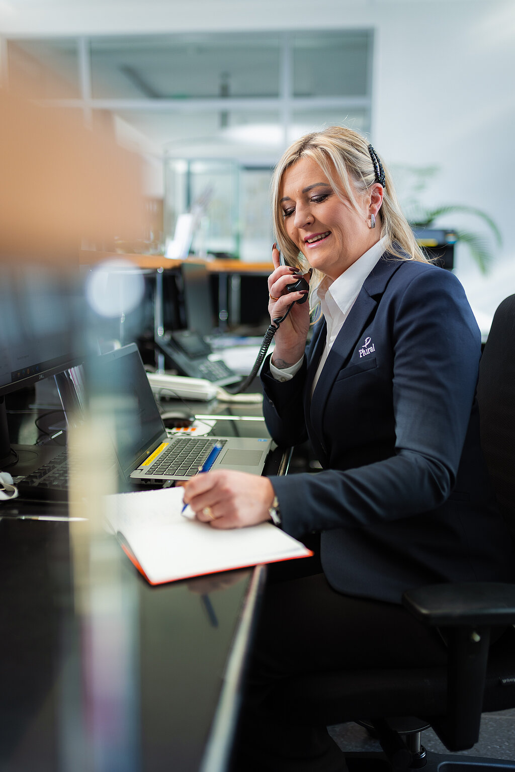 Eine junge blonde Dame, die am Empfang sitzt und telefoniert. Sie trägt eine weiße Bluse und einen Blazer.
