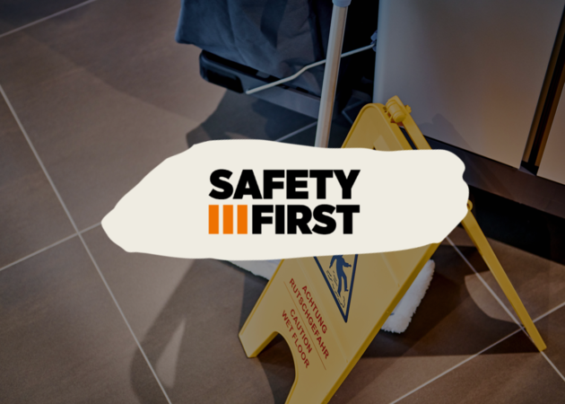 Ein gelbes Schild mit Achtung Rutschgefahr, davor das "Safety First" Logo der Compass Group.