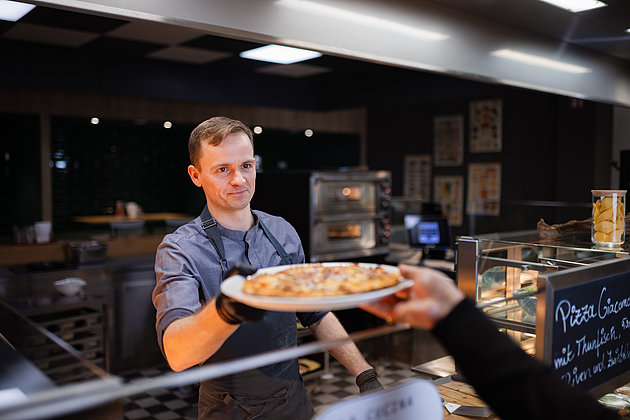 Ein junger Koch, der einem Gast eine Pizza über die Ausgabestelle gibt.