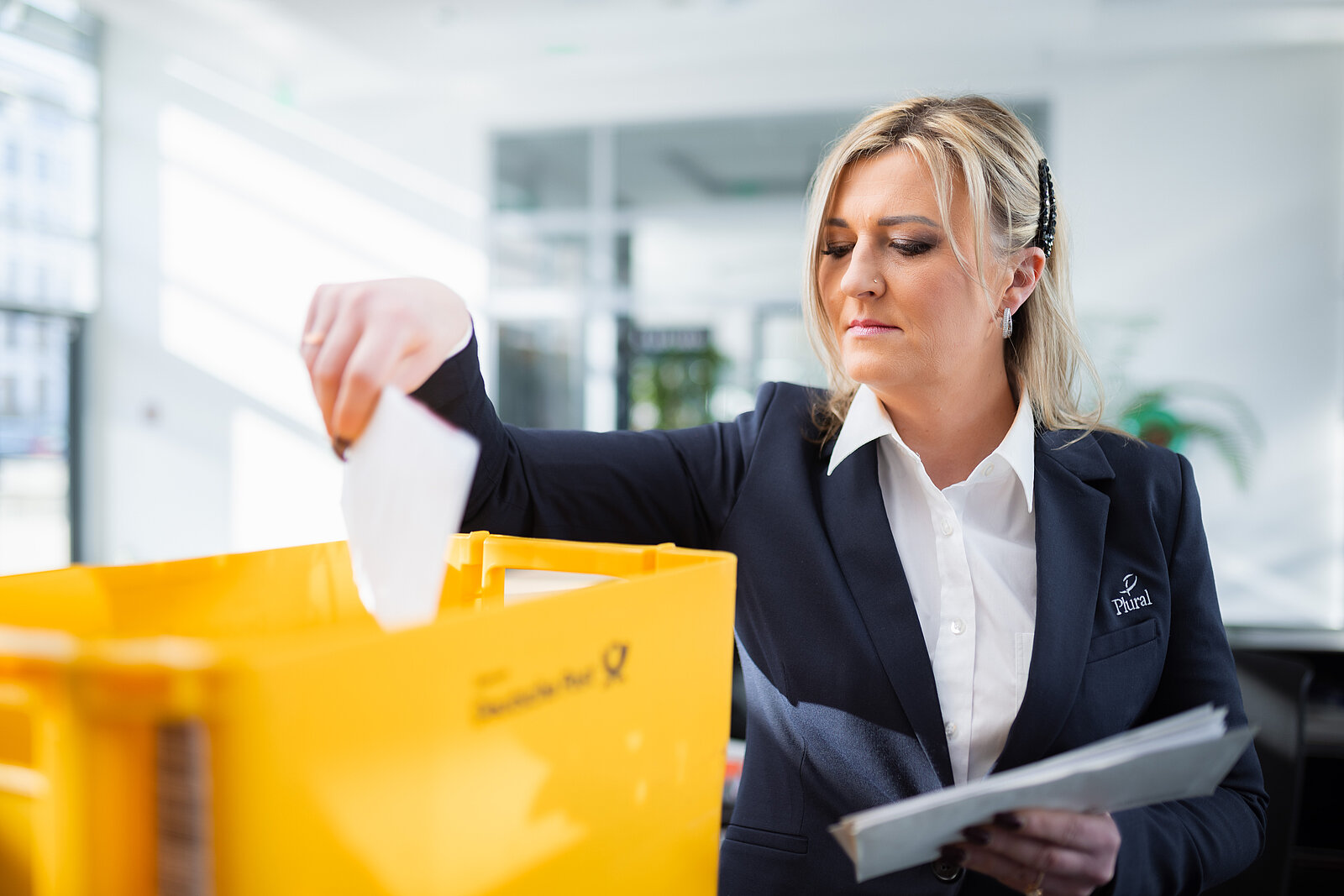 Eine junge Dame, die in eine Postbox Briefe einsortiert. Sie arbeitet am Empfang eines Unternehmens und trägt einen dunkelblauen Blazer.