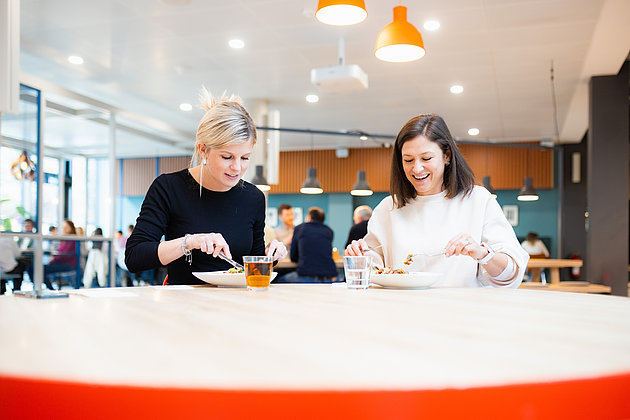 Zwei junge Frauen, die in einem schönen Ambiente in einer Betriebskantine ihr Mittagessen zu sich nehmen.