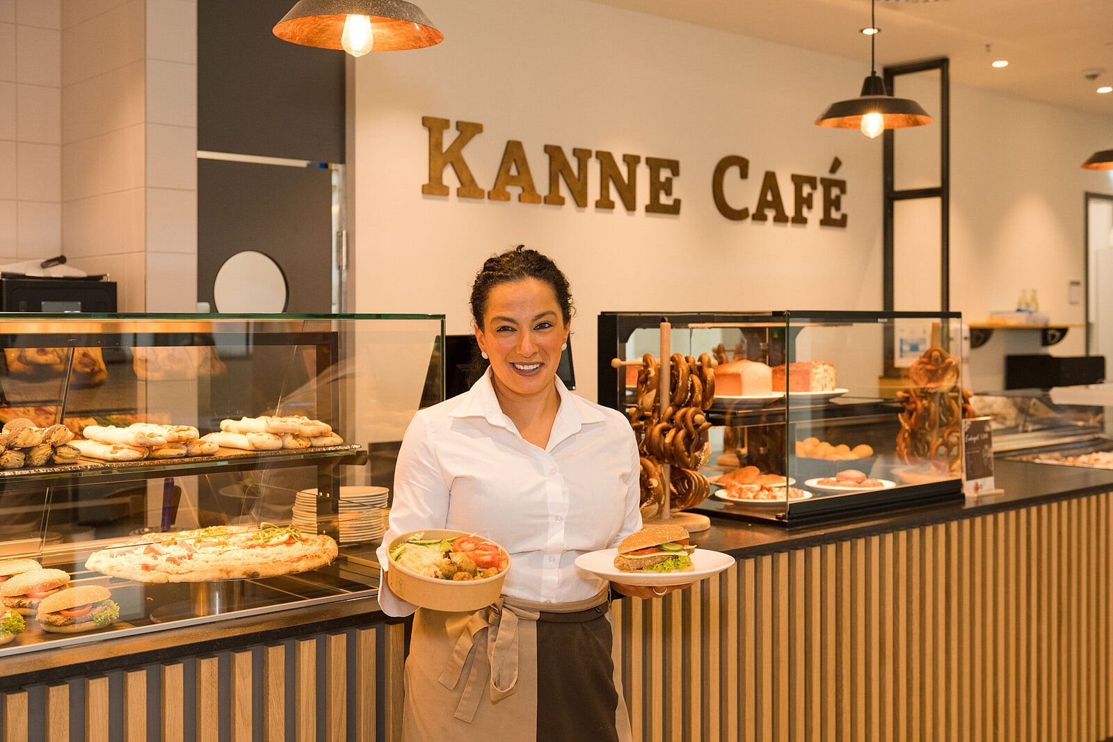Eine Frau, die vor einer Theke eines Kanne Cafe steht und in einer Hand einen Burger und in der anderen eine Bowl hält. Im Hintergrund ist der Schriftzug "Kanne Cafe" zu sehen.