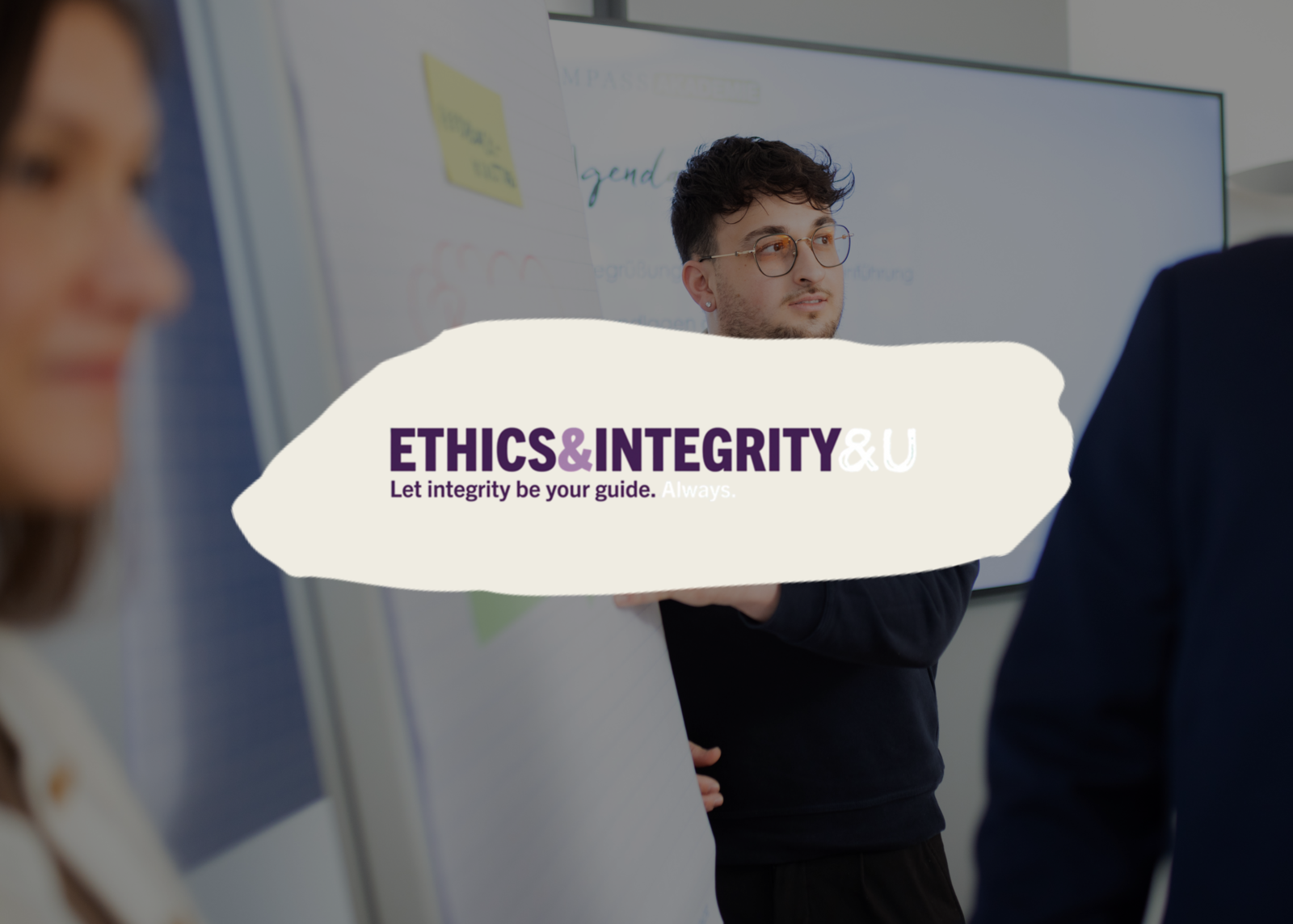Ein junger Mann, der an einem Flipchart steht und gerade etwas erklärt. Darüber ist das Logo der Compass Group Ethik und Integritätsinitiative "Ethics and Integrity" zu sehen.  