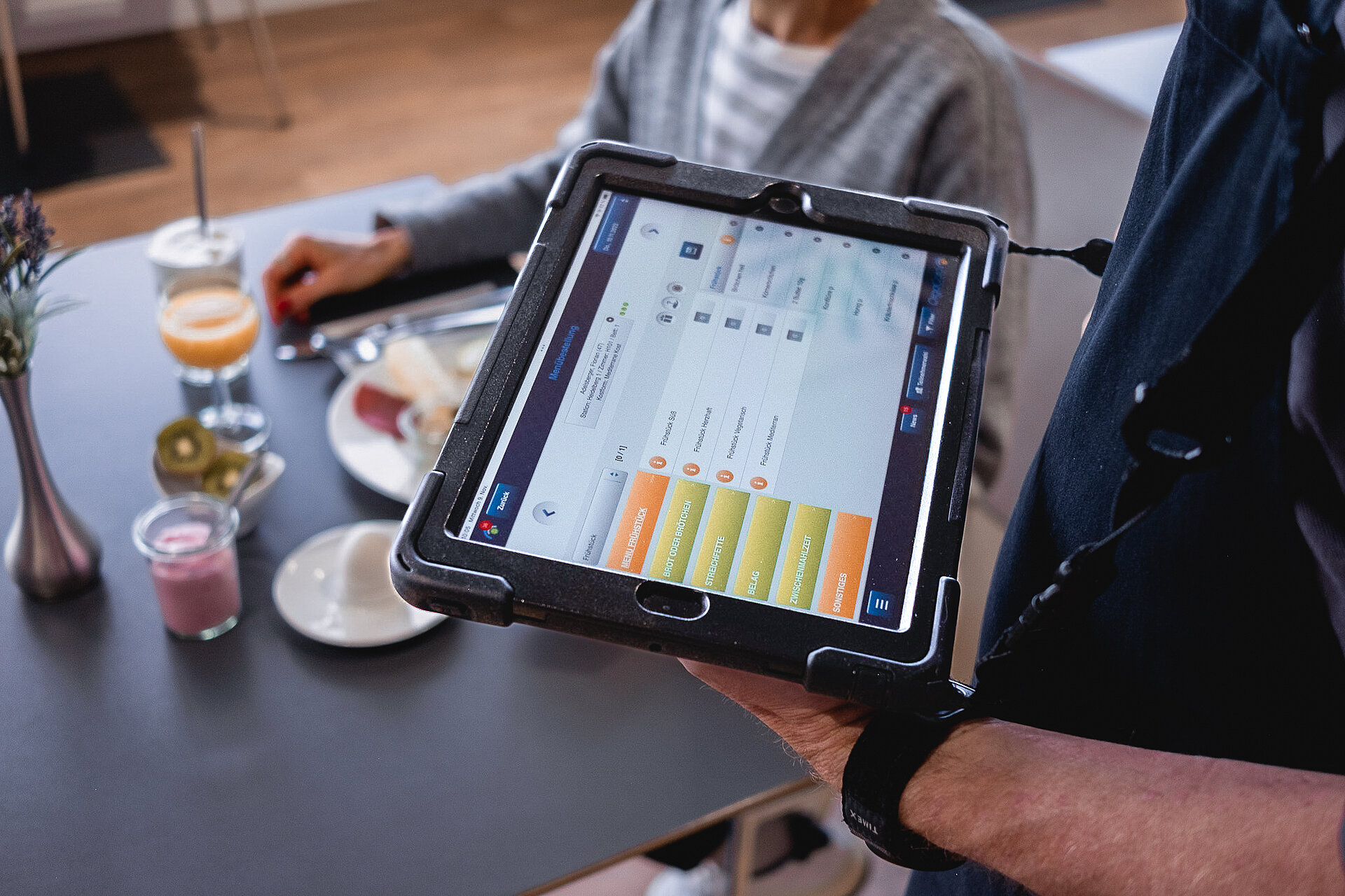 Ein Tablet mit einem Klinikmenüplan. Im Hintergrund ist eine Patientin an einem Tisch zu sehen.