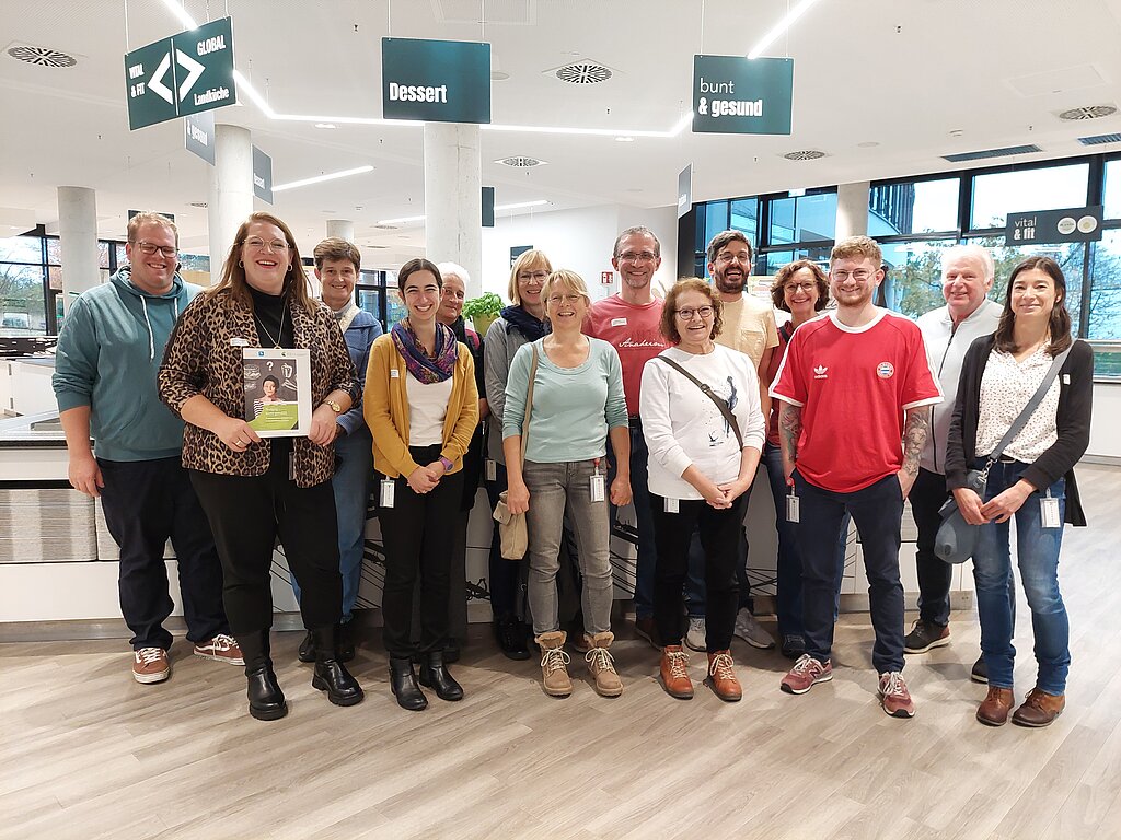 Eine Gruppe von Personen, die in einer Betriebskantine steht. Die Personen haben eine Infoveranstaltung zum Thema "Nudging" vom Betriebsleitenden erhalten.