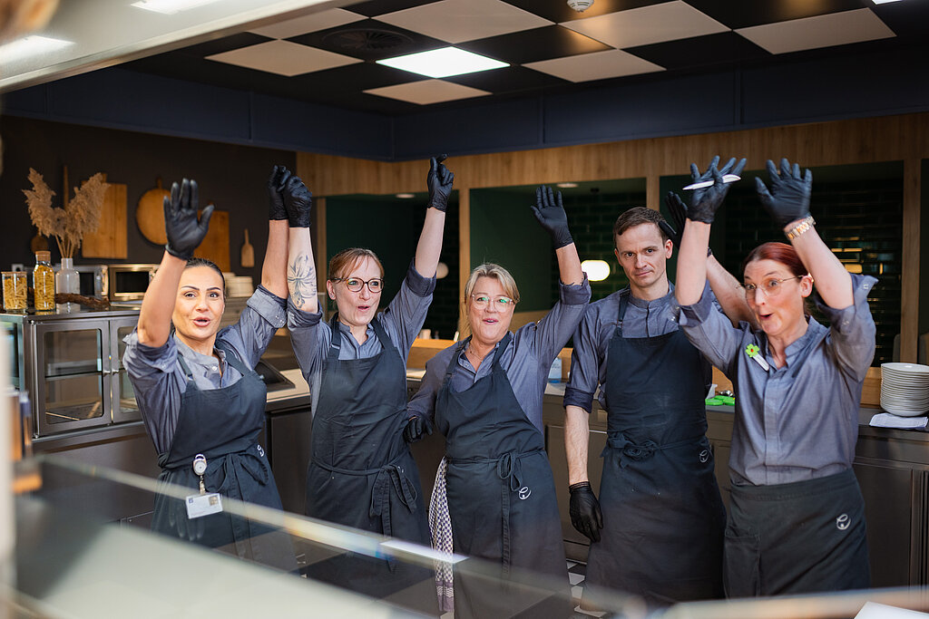 Ein Team bestehend aus Köchen und Küchenhilfen, das am Counter steht und die Hände in die Höhe schmeißt. Sie sind fröhlich und lachen.