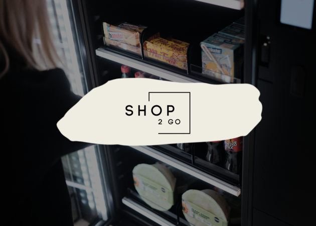 Shop2Go Eine junge Frau, die gerade Snacks aus einem Kühlschrank/Automaten holt. Davor ist das Logo der Compass Group Retailmarke Shop 2 Go zu sehen.