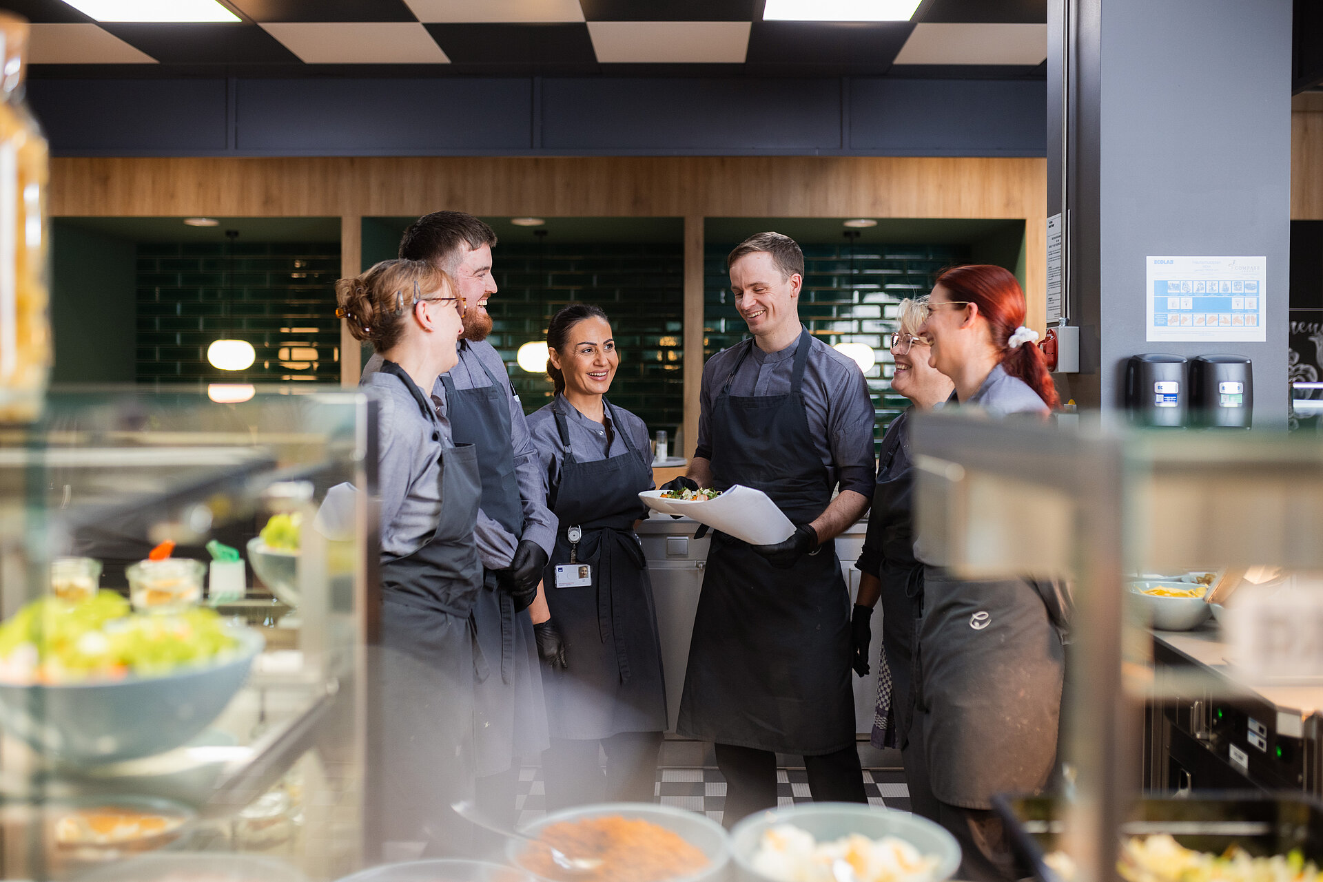 Ein Küchenteam in der Ausgabe einer Betriebsgastronomie. Der Betriebsleiter hält ein Gericht und eine Notiz in der Hand, er gibt seinen Kolleg:innen ein Briefing. Alle lachen dabei und sind gut gelaunt.