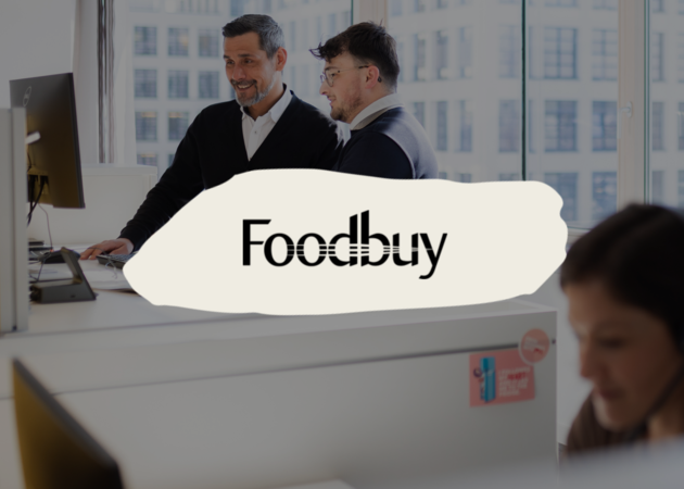 Foodbuy Zwei junge Männer, die an einem Computer im Büro stehen und über etwas sprechen, darüber das Logo der Compass Group Marke Foodbuy.