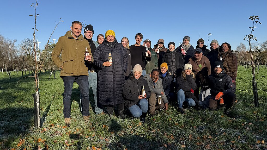 Eine Gruppe von Personen der Compass Group sowie von der Organisation Das Geld hängt an den Bäumen. Sie stehen bei Streuobstwiesen. Dahinter ein blauer Himmel.