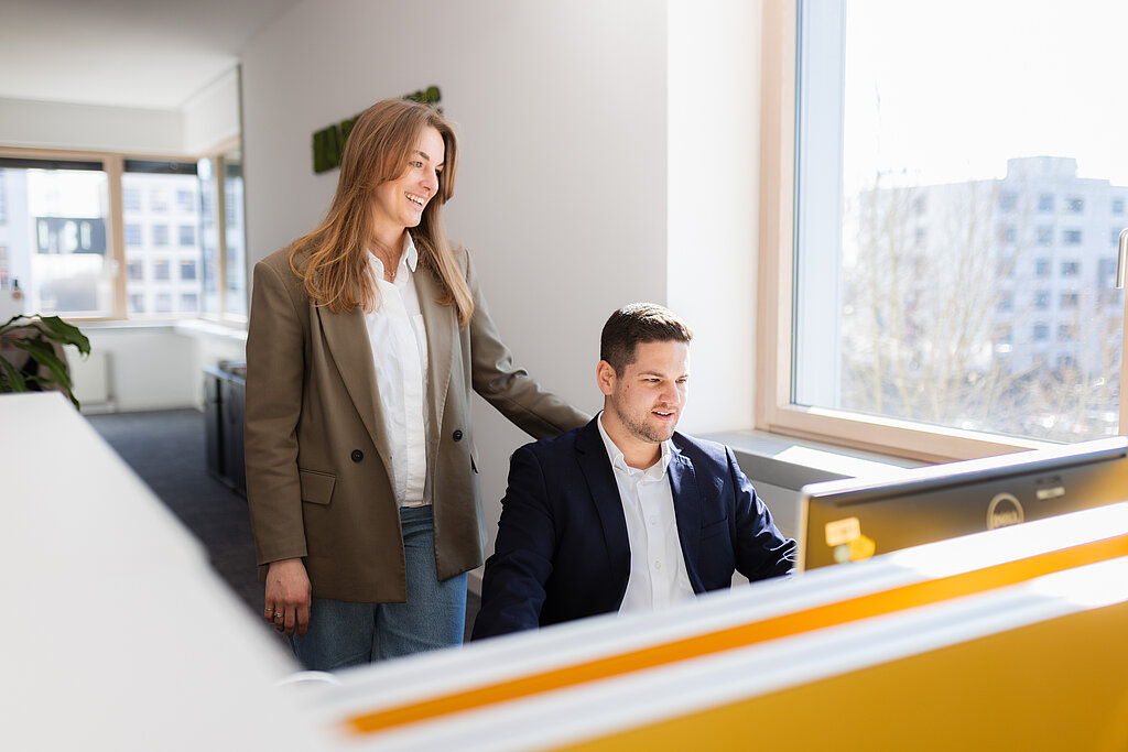 Eine junge Dame, die einem jungen Herren im Büro über die Schulter blickt. Der Herr sitzt und schaut in seinen Laptop, die Dame steht neben ihm und tut das ebenso.