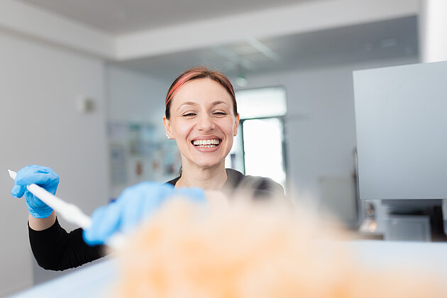 Multi Sektor Unternehmen Eine junge Reinigungskraft, die freundlich in die Kamera lächelt während sie mit einem Staubwedel eine Oberfläche reinigt.