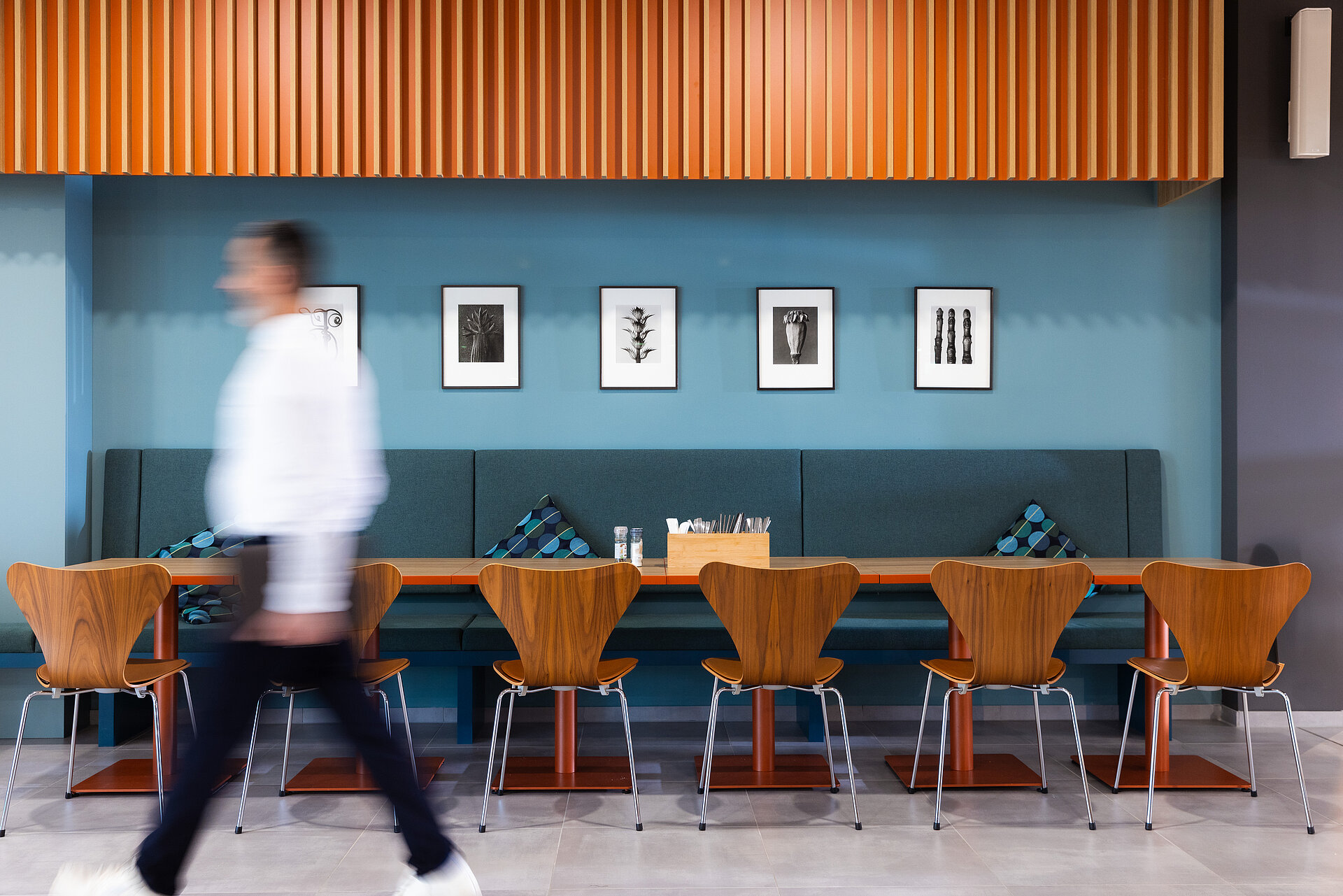 Ein Mann in schwarzer Hose und weißem Hemd, der in einem Betriebsrestaurant in einem schön gestalteten Gastraum geht. Die Wand vor der er geht, ist blau/grau.