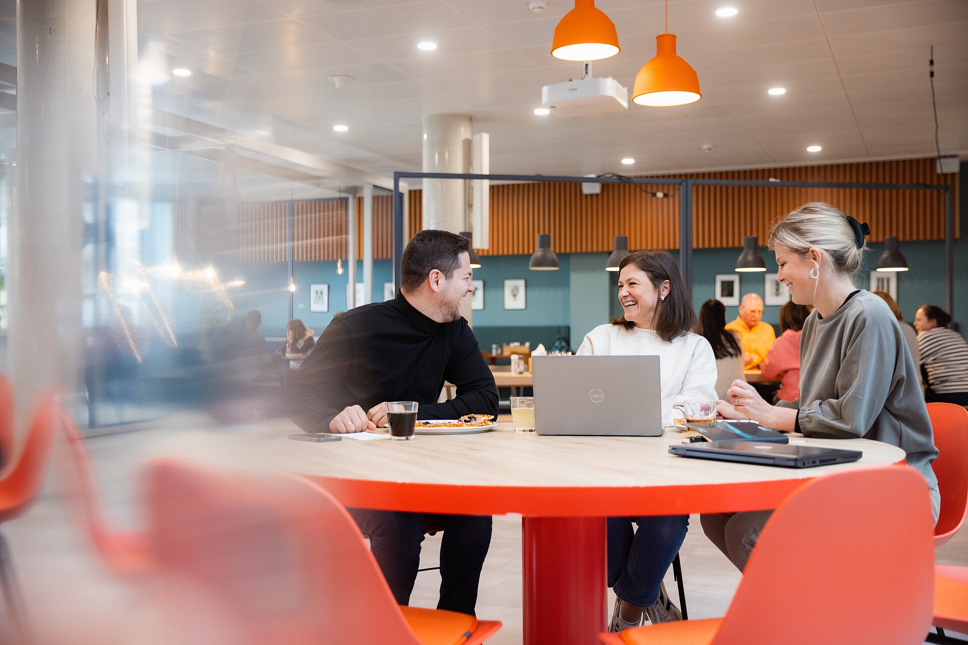 Drei junge Menschen, ein junger Mann und zwei junge Frauen, die in einer modernen Betriebskantine an einem Tisch sitzen und sich unterhalten. Sie scheinen dabei zu arbeiten.