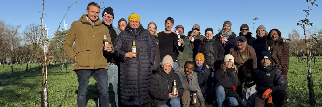 Eine Gruppe von Personen, die auf einer Streuobstwiese steht und freundlich in die Kamera lächelt. Zuvor haben die Personen Bäume eingepflanzt.