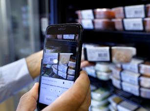 Scan & Go Eine Hand die ein Produkt aus einem Kühlregalnimmt und dieses dann mit dem Smartphone scannt.
